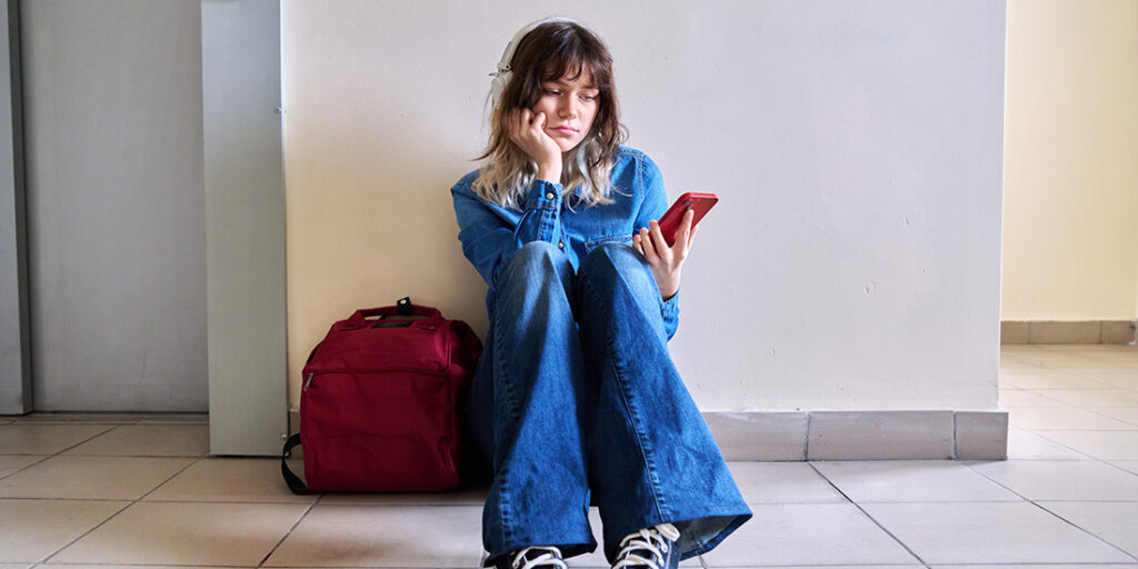 Sad young female student with backpack smartphone sitting near elevator on floor. Lonely frustrated teenage female, frustration problem pain depression concept