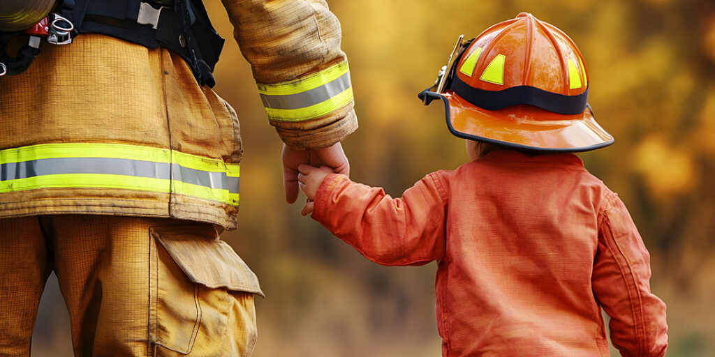 dirty firefighter holding hand of child in firefighter outfit, walking away from viewer