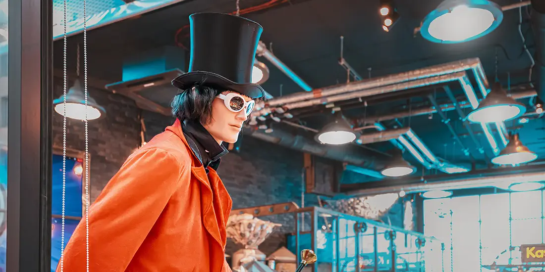 Person dressed in a whimsical costume resembling the fictional chocolatier Willy Wonka, wearing a bright orange coat, tall black top hat, and white round goggles. The person stands in an industrial-style interior with exposed pipes, circular hanging lights, dark brick walls, and large windows illuminated by blue and warm-toned lighting.