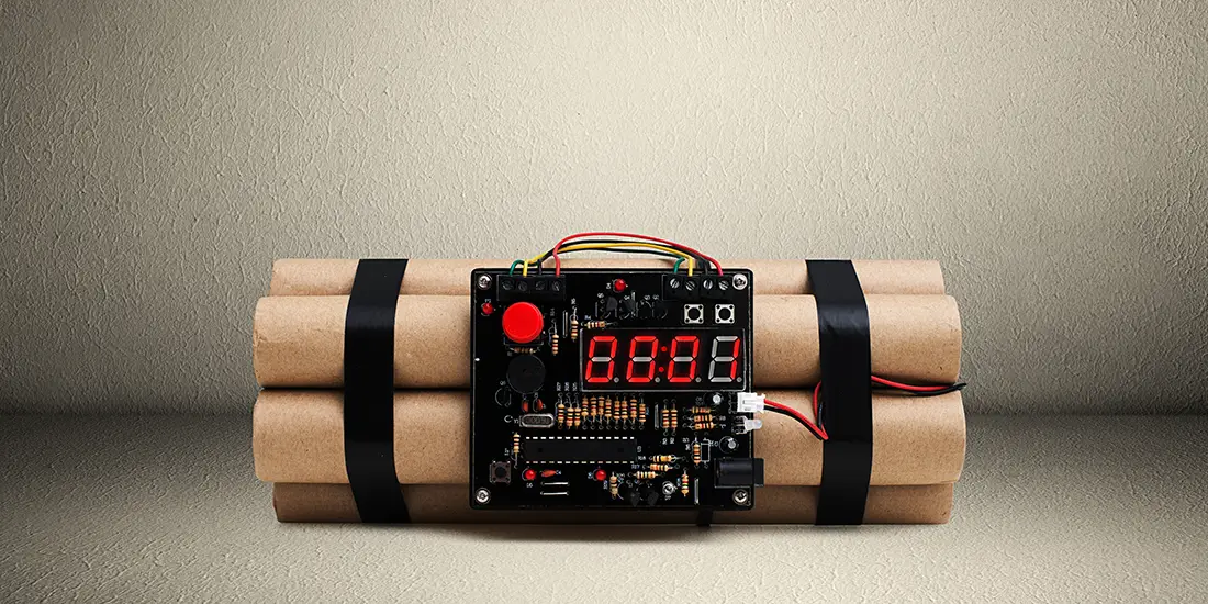 Improvised explosive device resembling a bundle of cylindrical tubes strapped together with black bands and connected to an electronic timer circuit board displaying red digital numbers. The device is placed on a flat surface against a plain textured wall background.