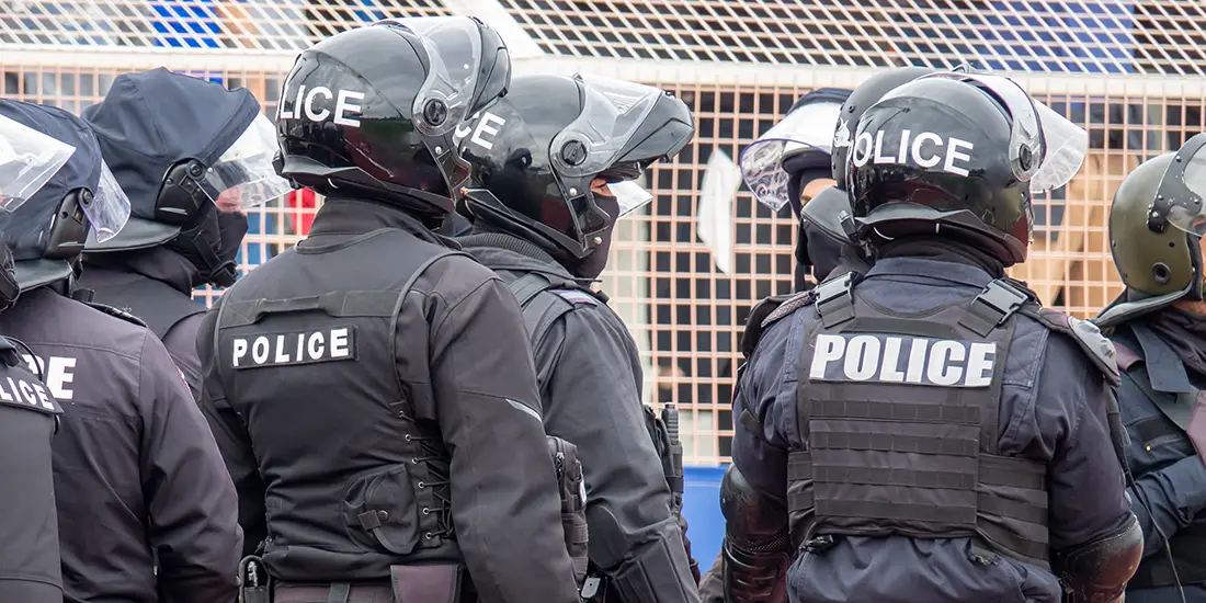 A group of police officers wearing black protective uniforms, body armor, and helmets with face shields stand closely together in front of a metal security fence. The word “POLICE” is visible on several helmets and uniform panels.