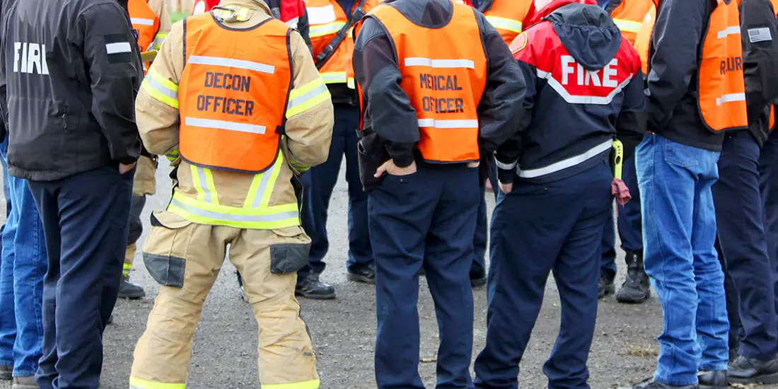 Emergency response personnel wearing high-visibility orange vests gather outdoors. Some vests are labeled “Decon Officer,” “Medical Officer,” and “Fire.”